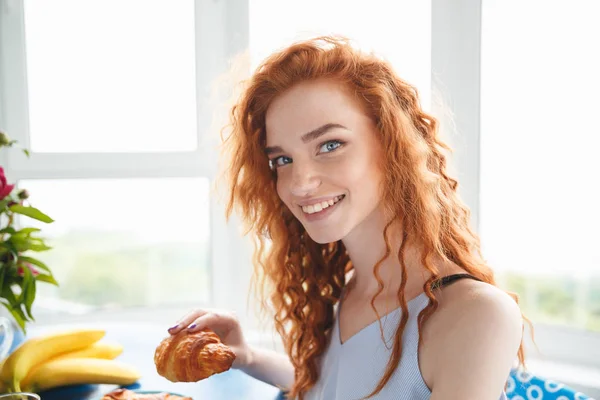 快乐的年轻红发女士坐在桌前吃牛角面包 — 图库照片