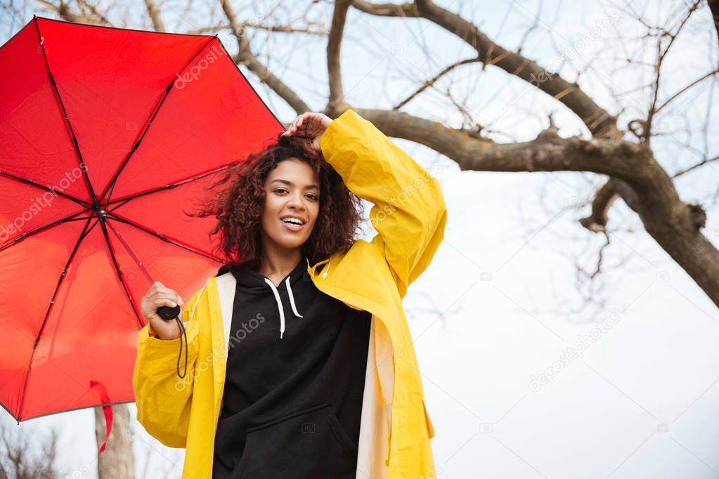 Happy african curly young lady wearing yellow coat with umbrella