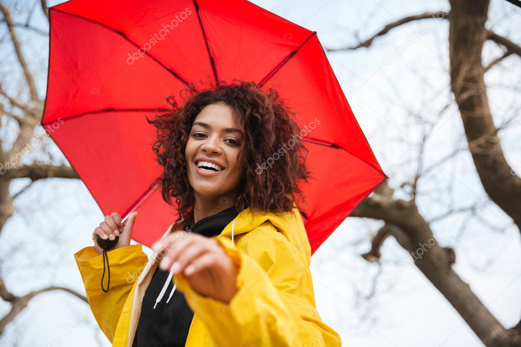 Happy african curly young lady wearing yellow coat with umbrella