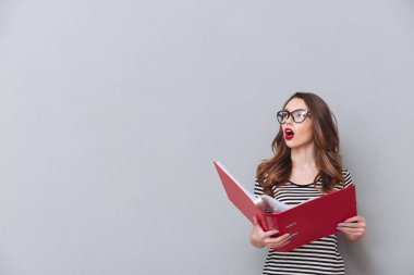 Gri duvardan ayakta ve klasör tutan düşünme genç bayan.