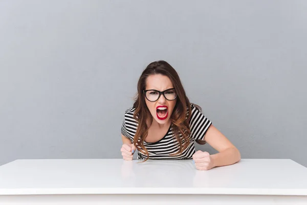 Wütend schreiende junge Frau mit Brille sitzt über grauer Wand. — Stockfoto