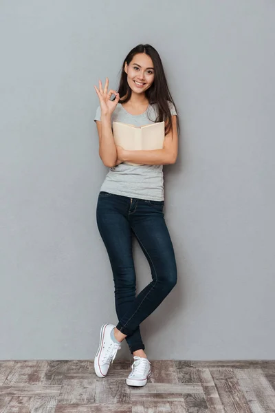 Sonriendo linda mujer joven sosteniendo libro y mostrando gesto ok — Foto de Stock