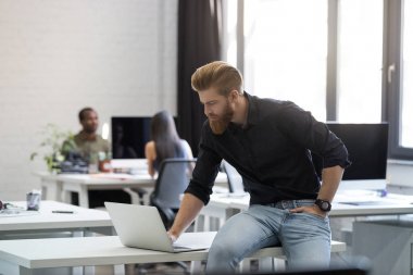 Genç adam, masanın üstüne oturan ve dizüstü bilgisayar ekranına bakarak