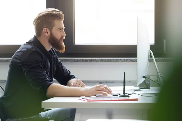 Sidovy av en ung skäggig man sitter vid sitt skrivbord — Stockfoto