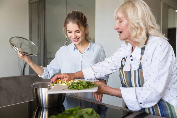 Lachende grootmoeder groenten in een pot zetten tijdens het koken — Stockfoto