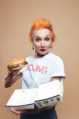 Olgun Kızıl saçlı kadın moda hamburger yeme. Görünümlü kamera.