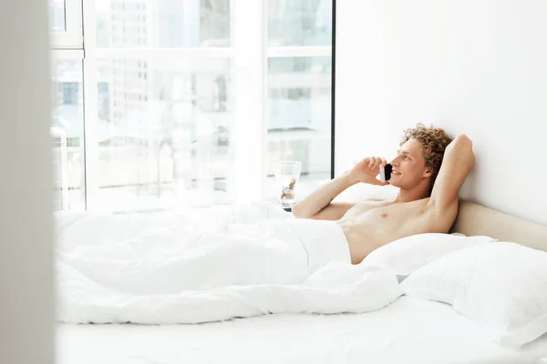 Веселий чоловік лежить в ліжку вдома, розмовляючи по телефону — стокове фото
