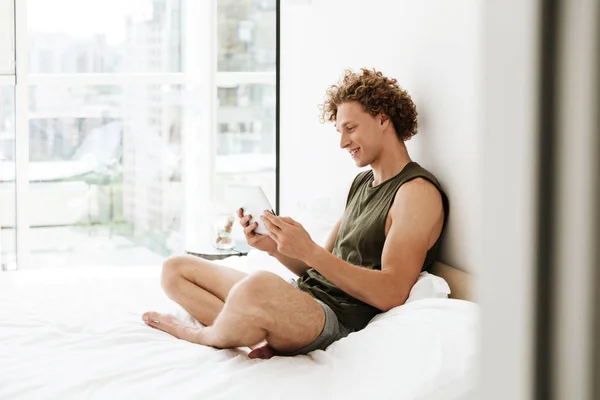 Щасливий чоловік сидить на ліжку вдома за допомогою планшетного комп'ютера — стокове фото
