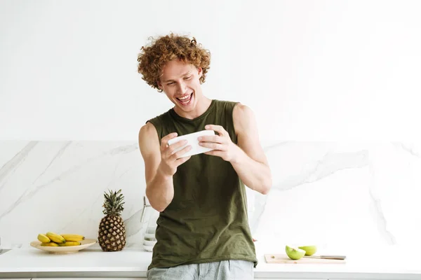 Lycklig man står på i köket och spela spel via telefon. — Stockfoto