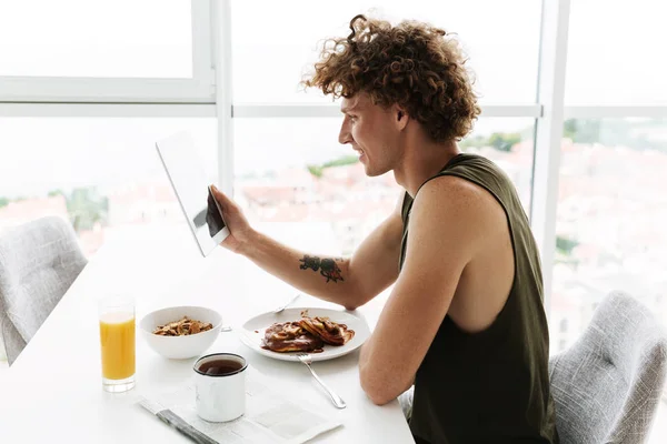 Bonito homem feliz sentado na cozinha enquanto usa tablet — Fotografia de Stock