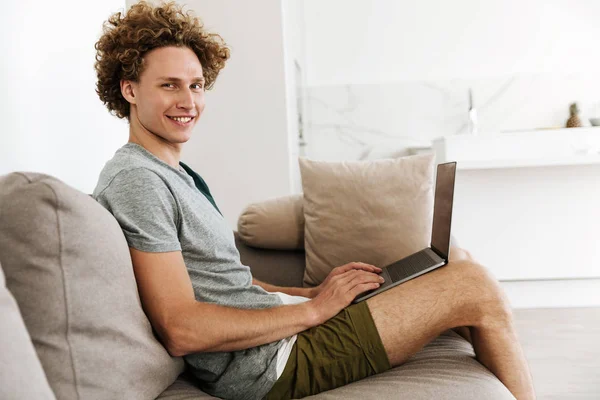 Dizüstü bilgisayar kullanan kanepe oturma yakışıklı neşeli adam. — Stok fotoğraf