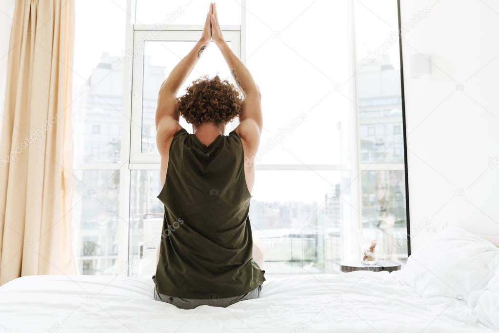 Back view image of curly man sitting on bed stretching.