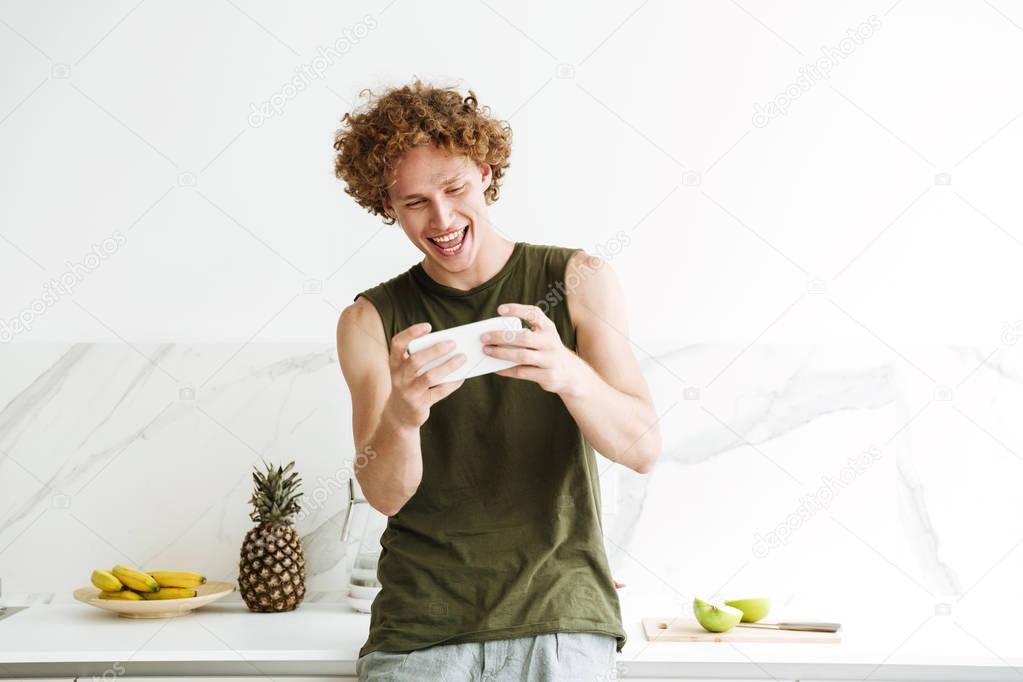 Happy man standing at the kitchen and play games by phone.
