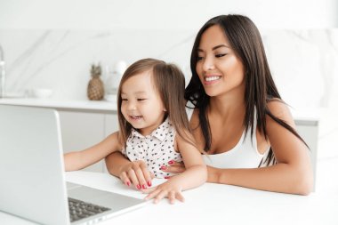 Gülümseyen Asya anne küçük kızıyla birlikte masada oturan ve dizüstü birlikte evde kullanma