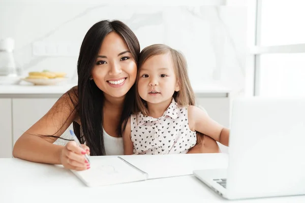 Glückliche junge Mutter mit kleiner Tochter mit Laptop Notizen schreiben — Stockfoto