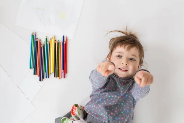 Улыбающаяся маленькая девочка лежит рядом с кучей цветных карандашей — стоковое фото