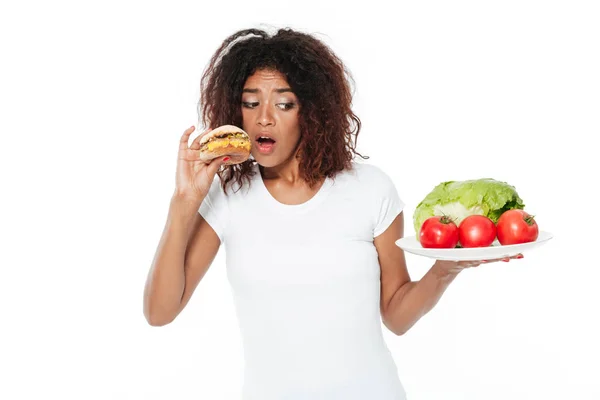 Confusa giovane donna africana che sceglie tra hamburger e verdure . — Foto Stock