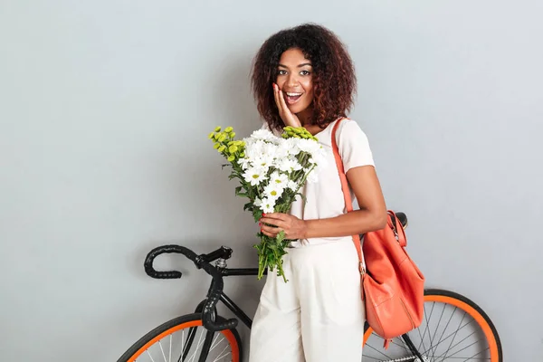 Улыбающаяся удивленная африканская женщина позирует с велосипедом, рюкзаком и цветами — стоковое фото