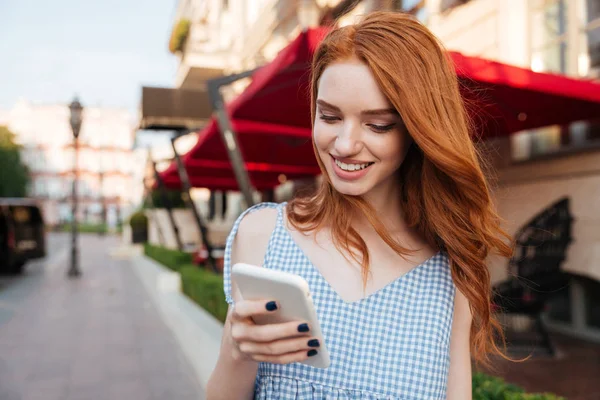 Веселая рыжая девушка с длинными волосами с помощью мобильного телефона в то время как — стоковое фото