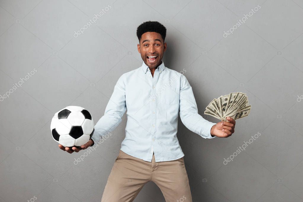 Happy smiling african man holding football and money banknotes isolated over gray background