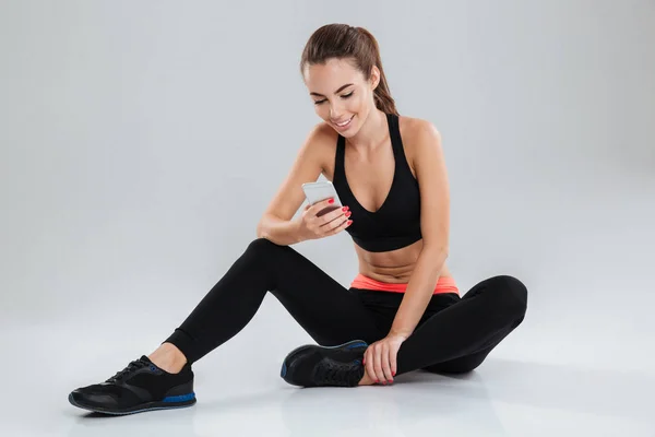 Χαμογελώντας γυμναστήριο γυναίκα κάθεται στο πάτωμα και χρησιμοποιώντας το smartphone — Φωτογραφία Αρχείου