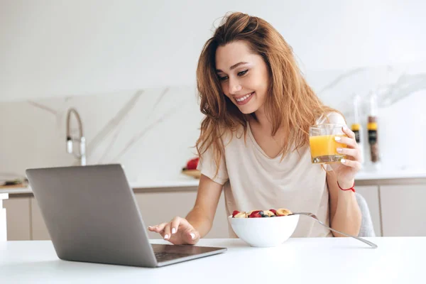 Mujer positiva con jugo usando el ordenador portátil en la cocina — Foto de Stock