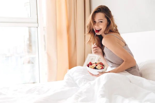 Wanita cantik ceria duduk di tempat tidur di dalam ruangan makan salad buah . — Stok Foto
