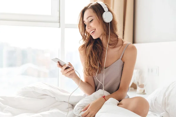 Dama sonriente en pijama escuchando música con smartphone y auriculares —  Fotos de Stock