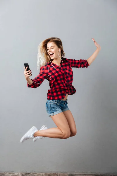 Glückliche junge blonde Frau springt und hört Musik per Telefon. — Stockfoto