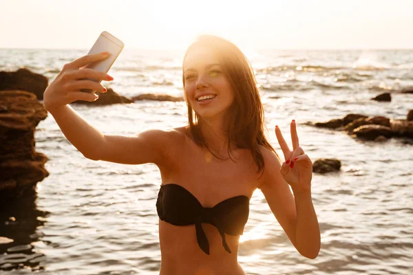 Joyful brunette woman in bikini making selfie on her smartphone — Stock Photo, Image