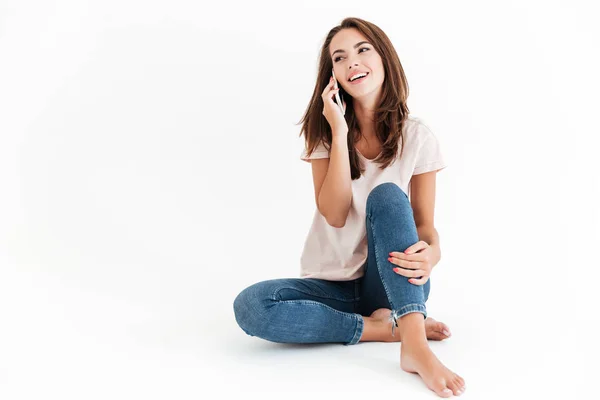 Smiling brunette woman sitting on floor and talking by smartphone — Stock Photo, Image