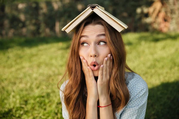 Grappige brunette vrouw zitten in park met boek op hoofd — Stockfoto