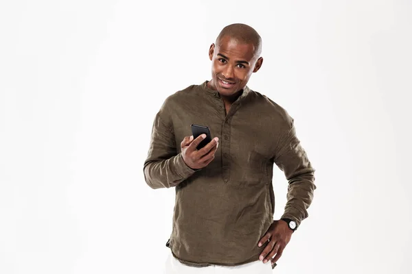 Улыбающийся африканский мужчина держит руку на бедре и использует смартфон — стоковое фото