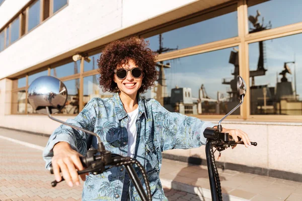 Imagem de mulher encaracolado feliz em óculos de sol sentado em moto — Fotografia de Stock