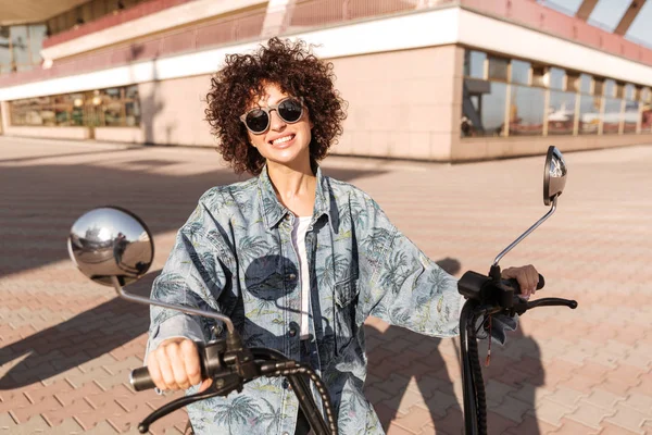 Image de joyeuse femme bouclée dans des lunettes de soleil assis sur la moto — Photo