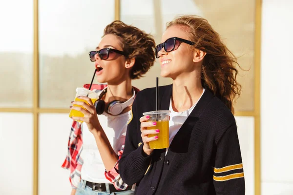 Två leende tonårsflickor i solglasögon dricka apelsinjuice — Stockfoto