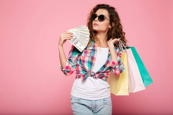Серьезная женщина в солнечных очках позирует с деньгами и сумками для покупок — стоковое фото