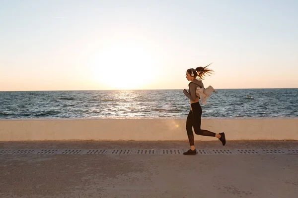 Νεαρή γυναίκα να ακούτε μουσική ενώ τρέχει στην παραλία στο ήλιο — Φωτογραφία Αρχείου