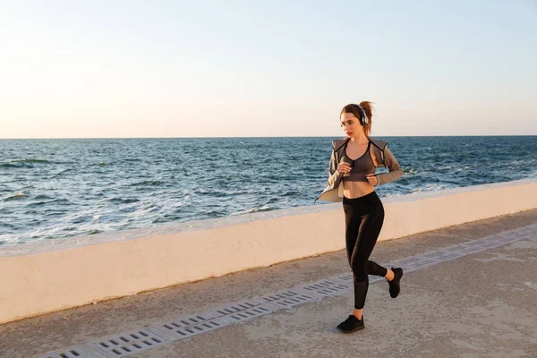 Bela mulher esportiva em fones de ouvido correndo à beira-mar — Fotografia de Stock