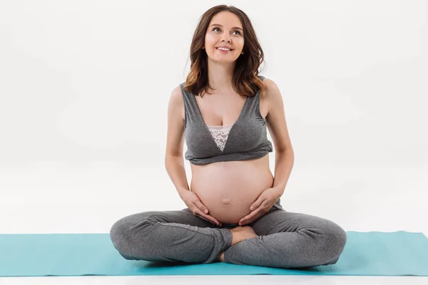 Smiling pregnant woman sitting on fitness mat and holding tummy — Stock Photo, Image
