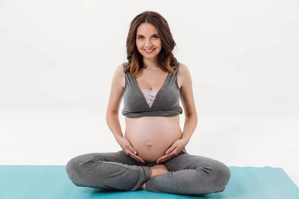 Smiling pregnant woman sitting on fitness mat and holding tummy — Stock Photo, Image