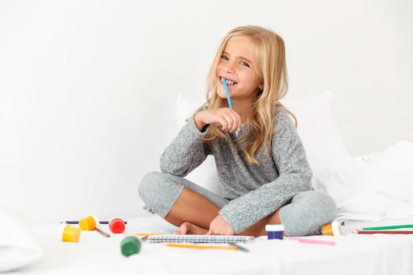 Menina loira feliz com lápis na boca sentado na cama, looki — Fotografia de Stock