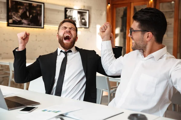 Dois homens de negócios felizes em ternos que levantam mãos perto de computador portátil, celebr — Fotografia de Stock