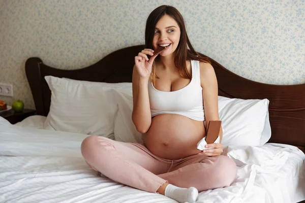 Gelukkig zwangere vrouw binnenshuis zittend op bed eten van chocolade — Stockfoto