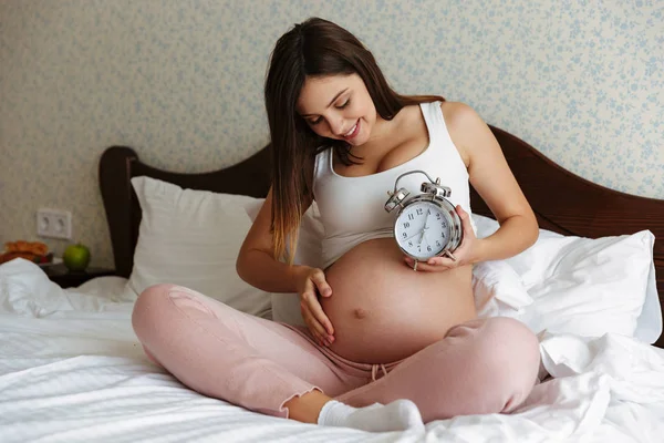 Vrolijke zwangere vrouw zittend op bed holding alarm — Stockfoto