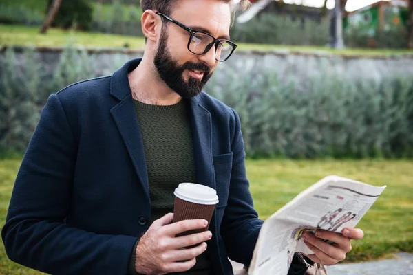Привлекательный молодой бородатый мужчина, сидящий на улице и читающий газету — стоковое фото