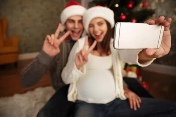 Sorrindo alegre mulher grávida com seu marido celebrando o Natal enquanto sentados juntos em um chão e tomando uma selfie em casa, casal mostrando gesto de vitória — Fotografia de Stock