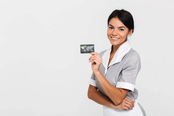 Alegre morena empregada doméstica no uniforme segurando cartão de crédito e olhar — Fotografia de Stock