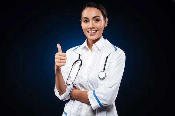 Mujer joven positiva médico con estetoscopio mostrando el pulgar hacia arriba —  Fotos de Stock