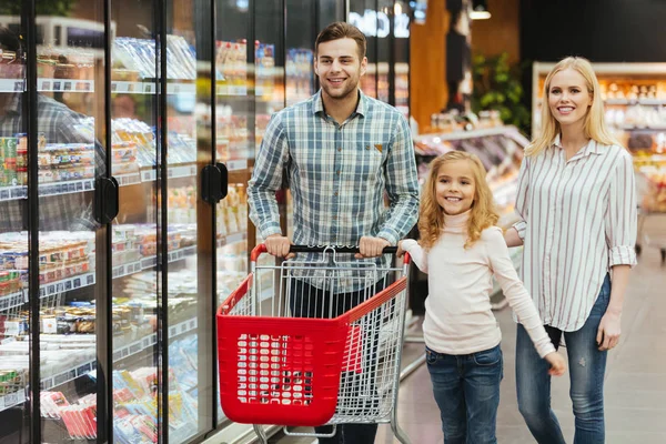 Glückliche junge Familie mit einem Kind, das mit einem Einkaufswagen unterwegs ist — Stockfoto